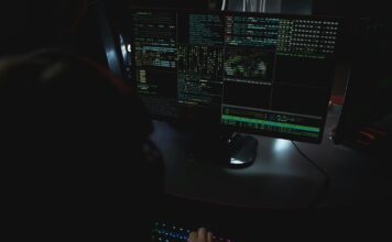 Anatomia DDoS od strony wydajności: jak przygotować infrastrukturę, by przetrwać atak Ciemne biuro z monitorami pokazującymi kod związany z cyberbezpieczeństwem