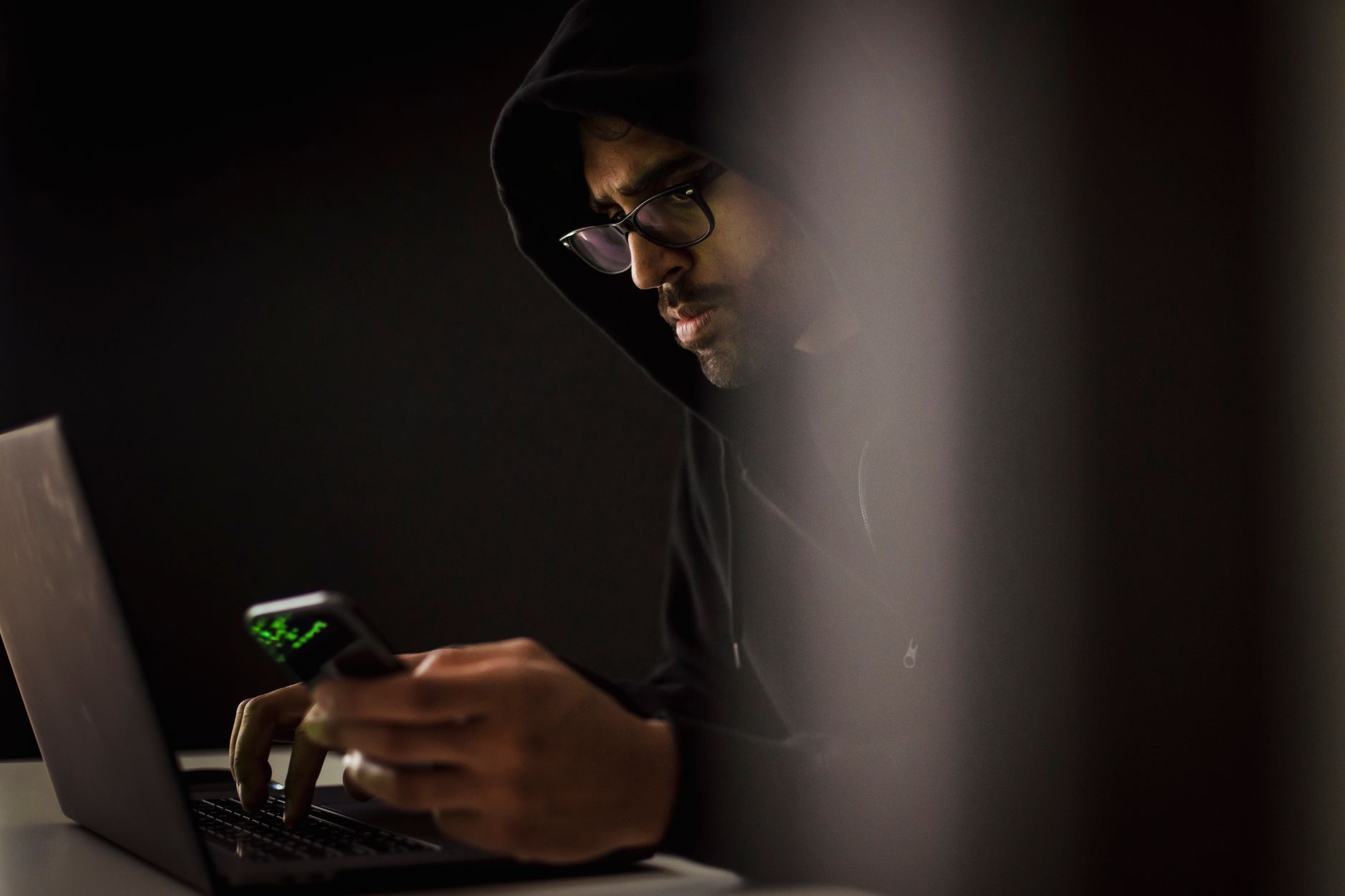 Brodaty mężczyzna w kapturze rozmawia przez telefon przy netbooku w ciemnym poko